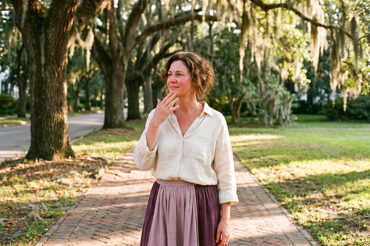 A woman pausing on an oak-lined sidewalk on a warm afternoon, fanning her face with her hand